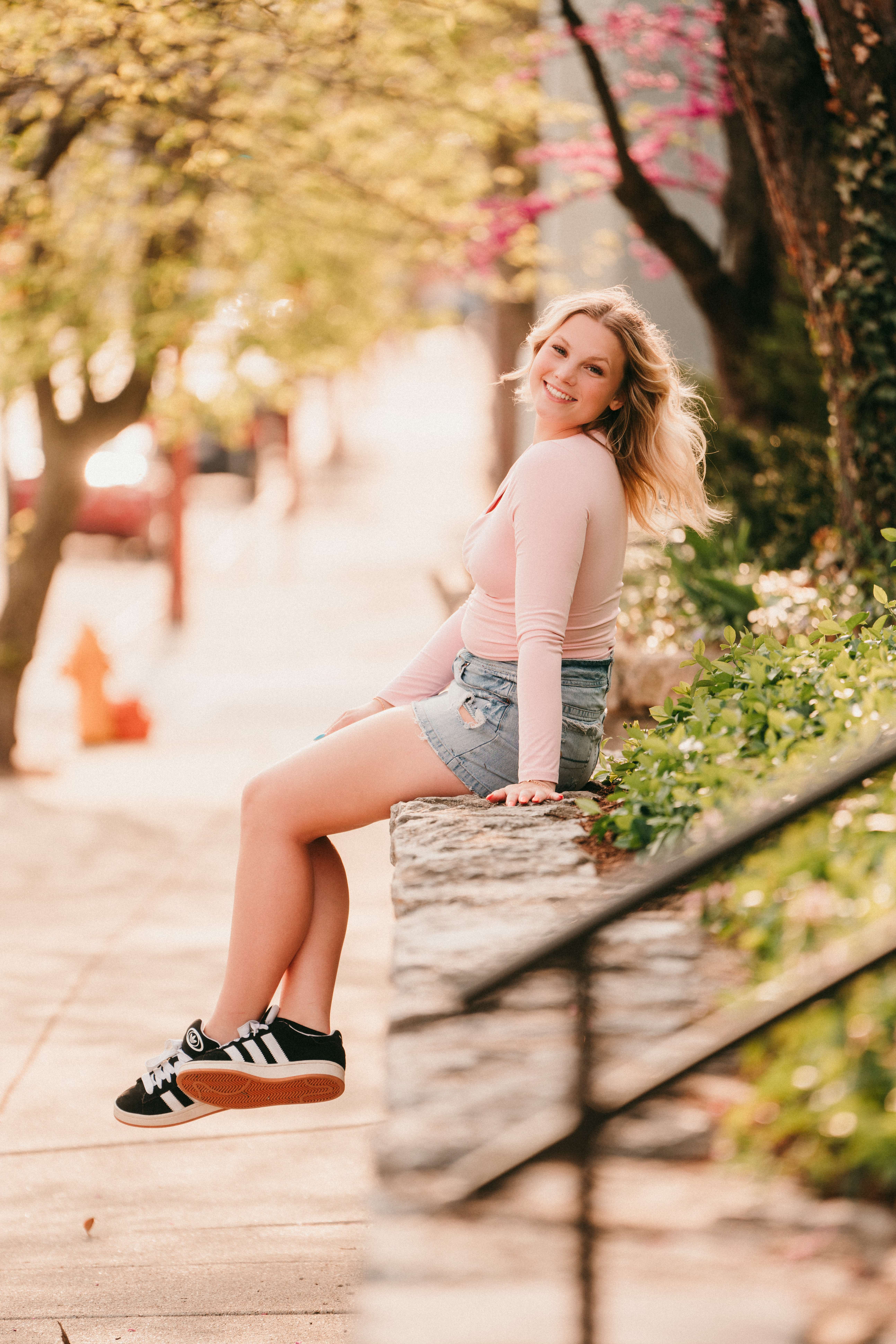 Blonde senior in casual attire smiling while sitting on a stone wall outdoors.