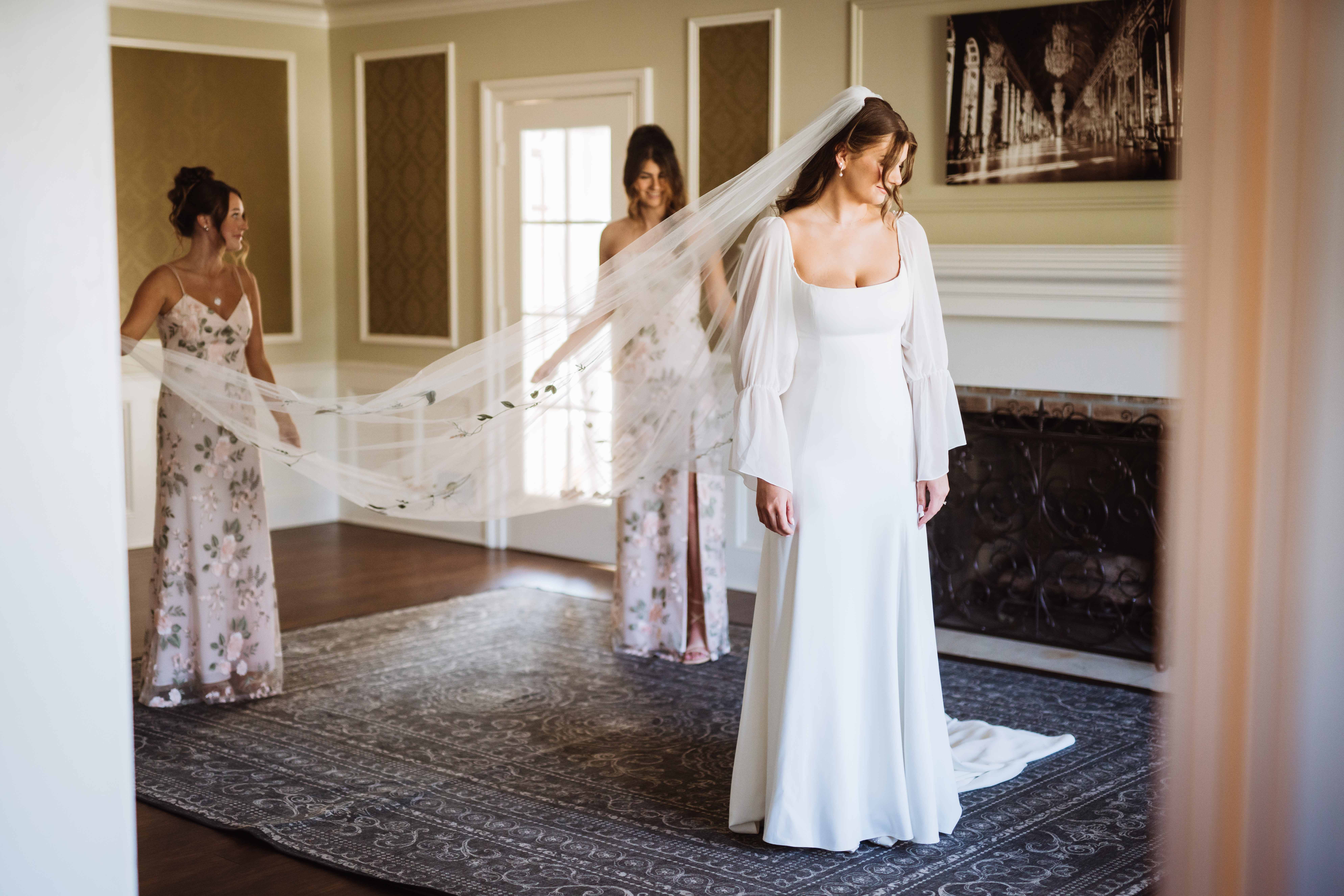 Bride having her long veil adjusted by bridesmaids in an elegant room.