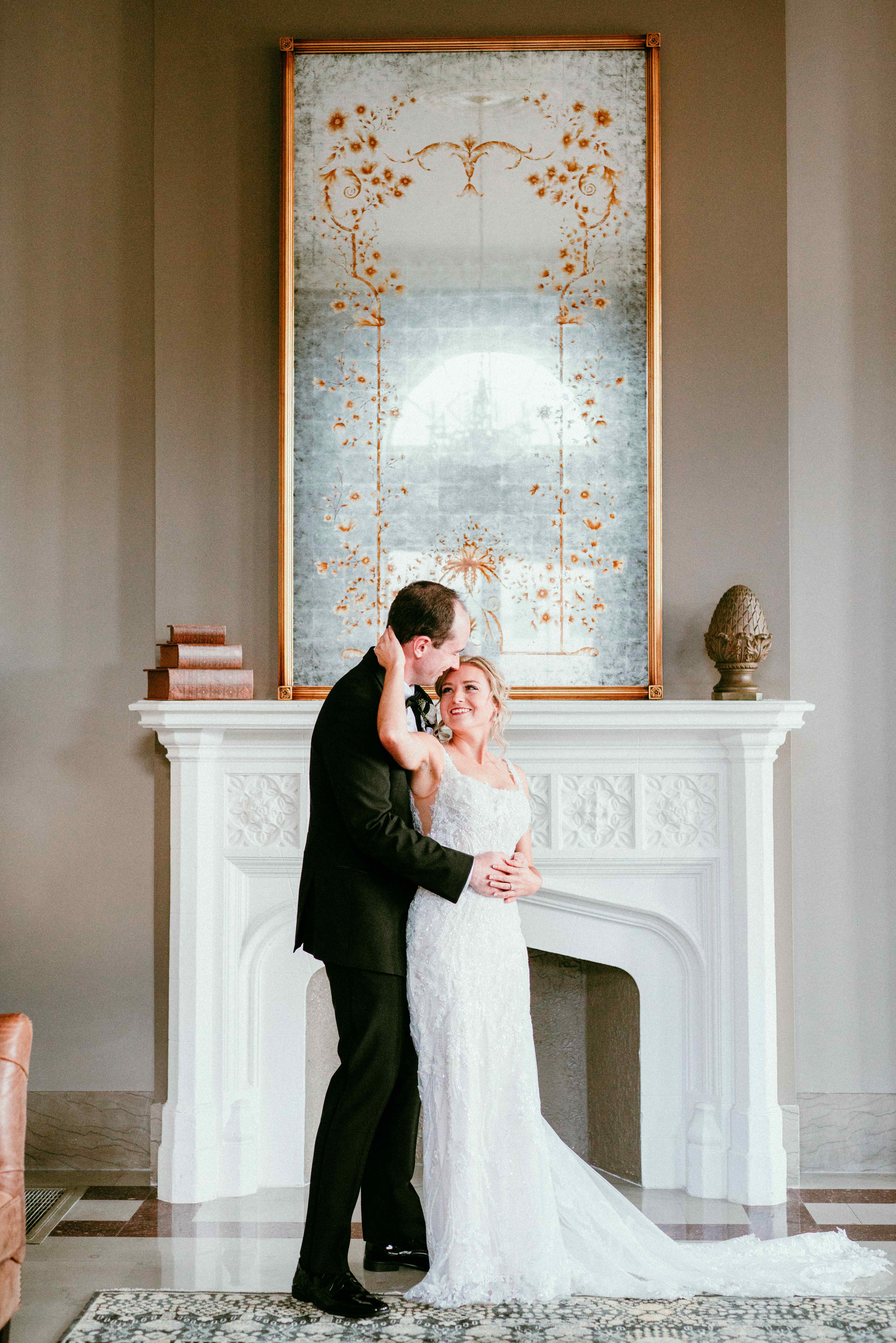 Groom kissing bride's temple as they embrace in front of an ornate fireplace.